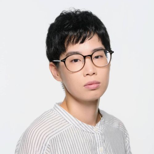 白とグレーのノーカラーストライプシャツに茶色の丸メガネでこちらを見る、山田ようのバストアップ写真。