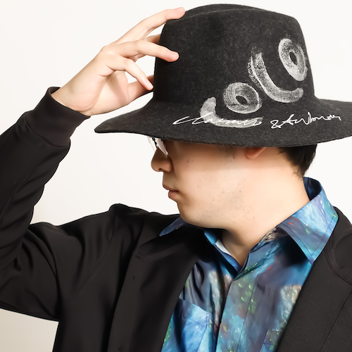 白い刺繍入りの帽子を右手で被り、黒いジャケットに黒と青のマーブル模様のシャツを着た石神哲朗の横顔の写真。