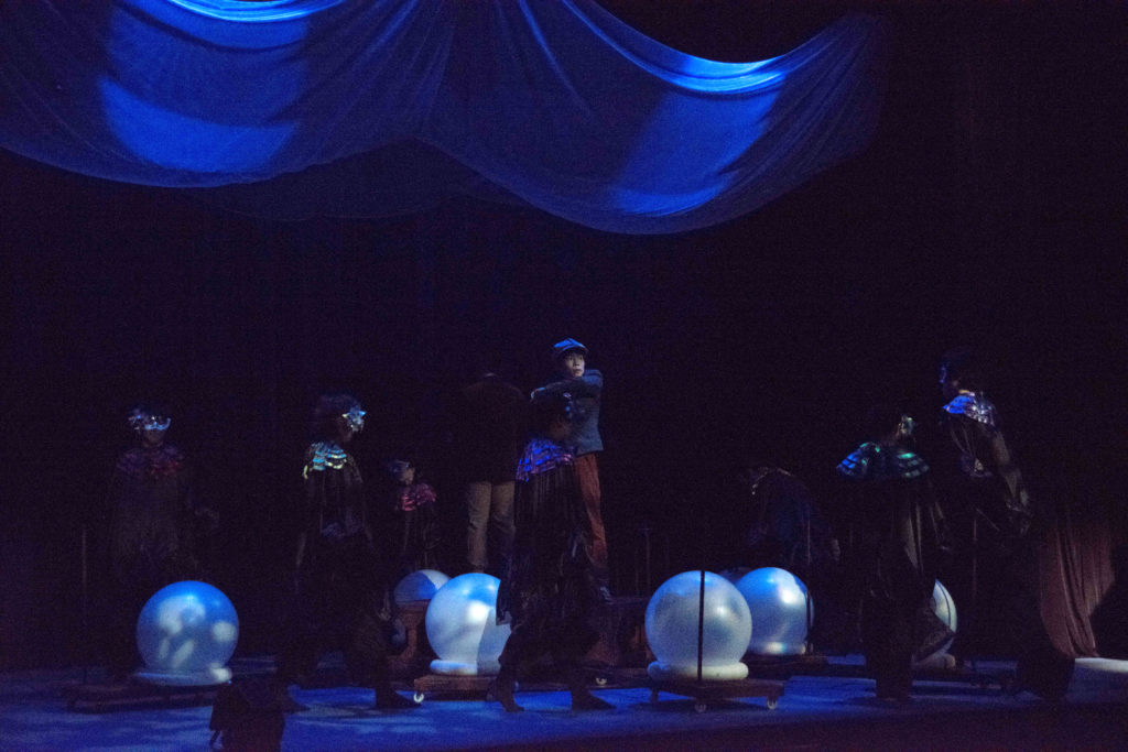 「銀河鉄道の夜」の舞台公演写真。舞台上には魂をイメージした７つの白い球体。７つの球の中心に立つジョバンニ役の男性。仮面をつけたコロスがジョバンニを囲んでいる。