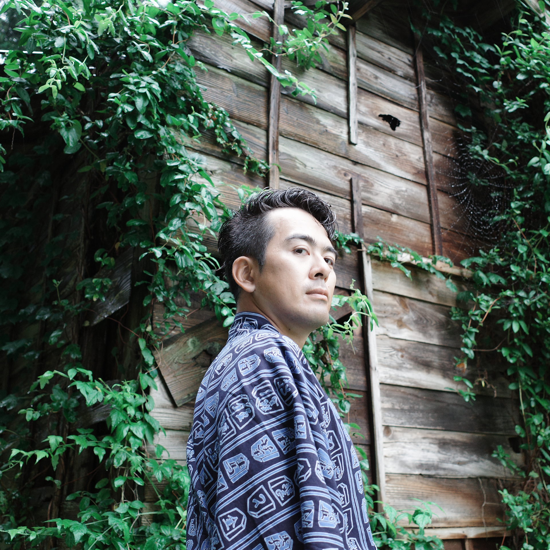 木造の小屋の前に立つすずきたいじ。将棋模様の浴衣姿。小屋の壁には鮮やかな緑の蔦。